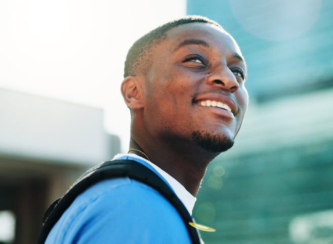 Photograph of a person wearing a blue shirt and black backpack standing outdoors with a modern building in the background. The image captures a casual, urban setting with natural lighting and a shallow depth of field.