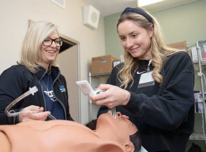 Photograph of two medical trainees practicing intubation on a medical mannequin in a clinical training room. One trainee holds a laryngoscope while the other operates a device, with medical supplies and equipment visible on shelves in the background.